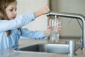 A young girl filling a clear glass with water from a sleek kitchen faucet, relying on a well water filtration system to ensure the water is clean and safe to drink.