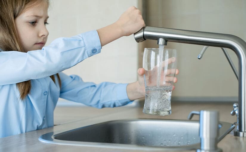 A young girl filling a clear glass with water from a sleek kitchen faucet, relying on a well water filtration system to ensure the water is clean and safe to drink.